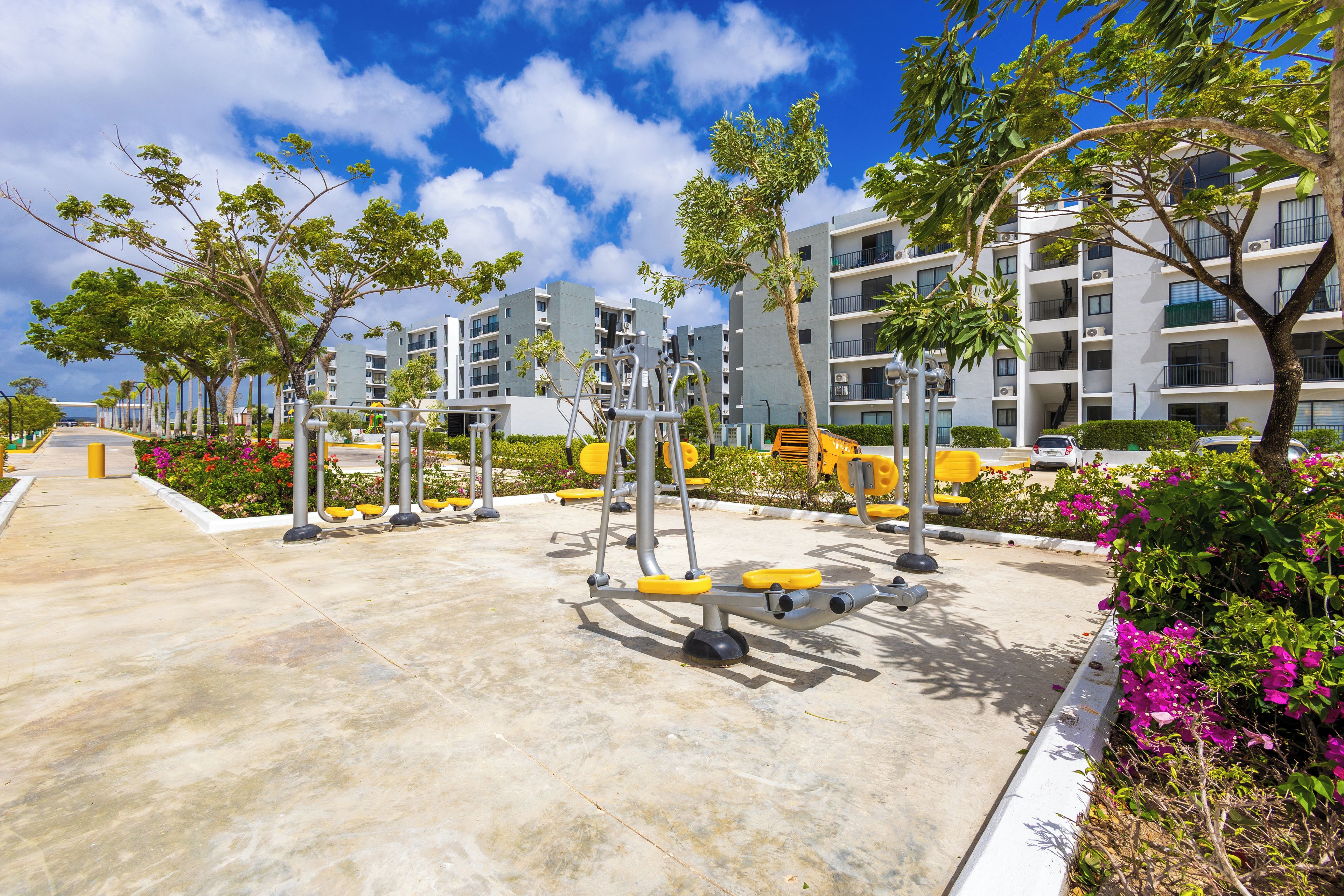 Un gimnasio con equipamiento de ejercicio, rodeado de vegetación y edificios de apartamentos modernos.