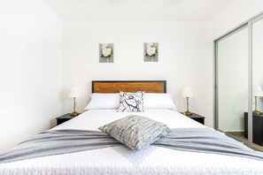 Ein ordentlich bezogenes Bett mit einem Holzkopfteil, zwei Nachttischlampen und zwei an der Wand hängende Blumenbilder.