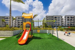 Ein Spielplatz mit Rutsche und Schaukeln, umgeben von Kunstgrashal und Palmen, mit Wohngebäuden im Hintergrund.