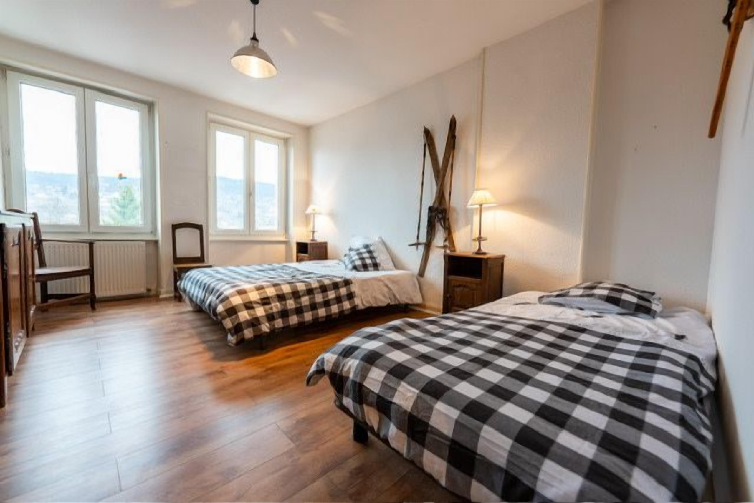 A bedroom with two beds, a desk, and a chair. There are windows and a view of the outdoors.