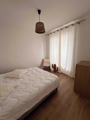Ein Schlafzimmer mit einem Bett, einem Sessel, einem Kaktus, einer Holzkommode und einem Fenster mit Vorhängen.