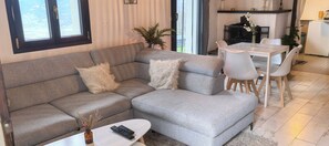 Una sala de estar con un sofá gris, dos almohadas mullidas, una mesita con un florero y controles remotos, y un comedor con una mesa y sillas blancas.