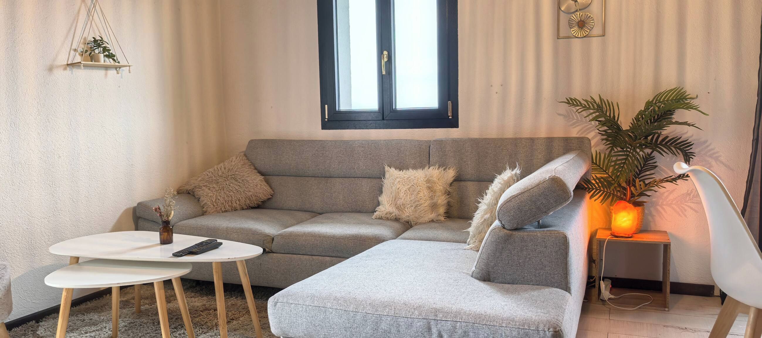 Una sala de estar con un sofá gris, una mesa de centro blanca y una mesita de noche con una lámpara.