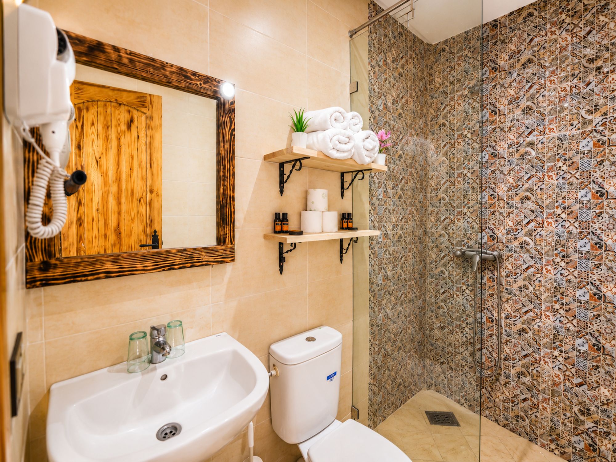 Un baño con puerta de madera, un lavabo, un inodoro, una ducha con pared de azulejos estampados y estantes con toallas y artículos de aseo.