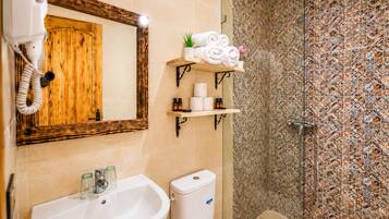 Un baño con puerta de madera, un lavabo, un inodoro, una ducha con pared de azulejos estampados y estantes con toallas y artículos de aseo.