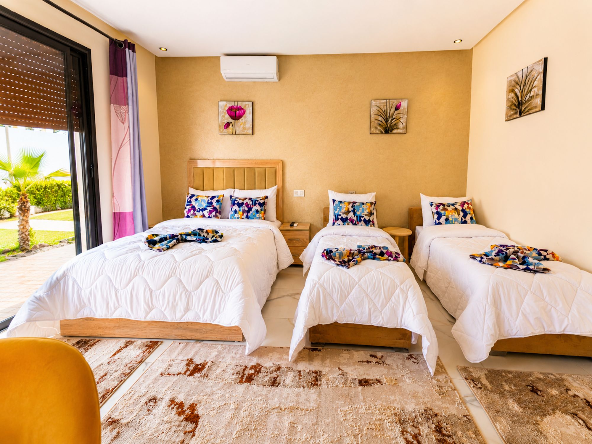 Habitación de hotel con tres camas, un ventanal con persianas, una mesita pequeña y una silla.