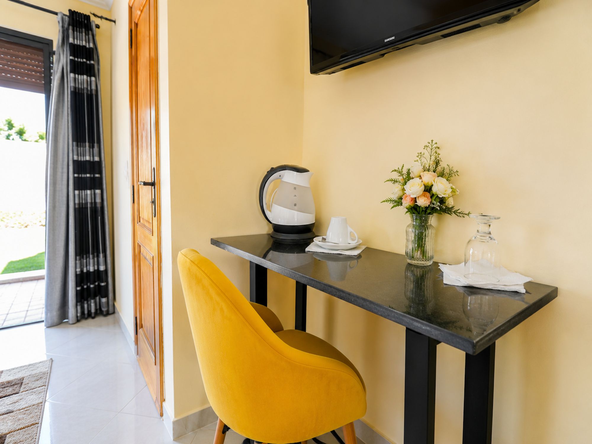 Ein modernes Zimmer mit einem gelben Sessel, einem Tisch mit Wasserkocher, einer Vase mit Blumen und einem flachbildfernseher an der Wand.