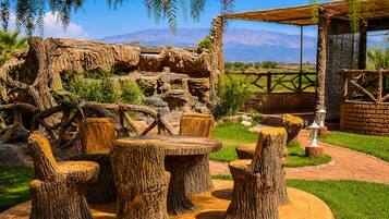 A rustic outdoor seating area with wooden tables and chairs, surrounded by a natural landscape with a view of distant mountains.