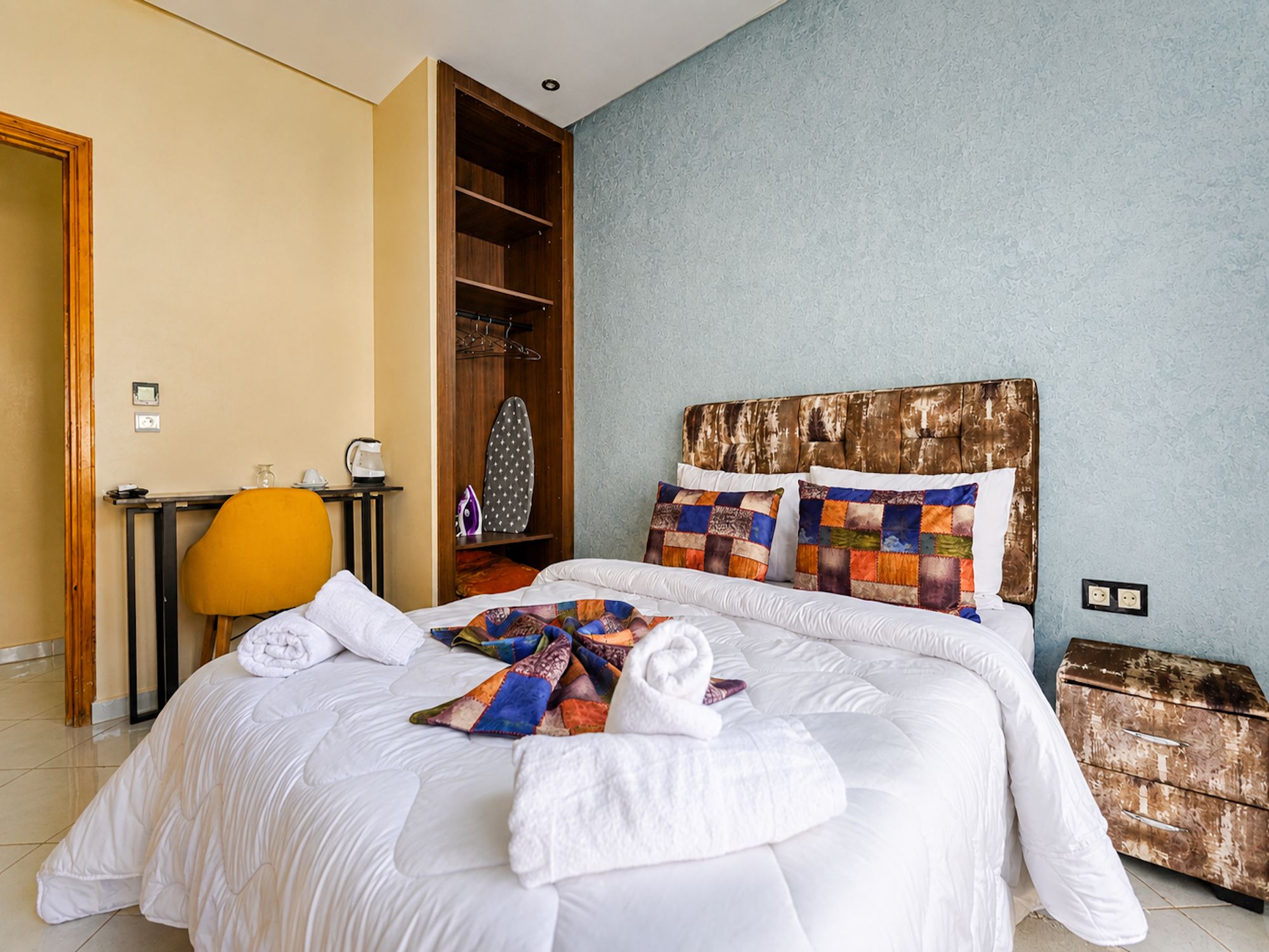 Ein Hotelzimmer mit einem großen Bett, einem Holz-Kopfstück, einem kleinen Tisch mit Wasserkocher, einem Sessel und einem Kleiderschrank.