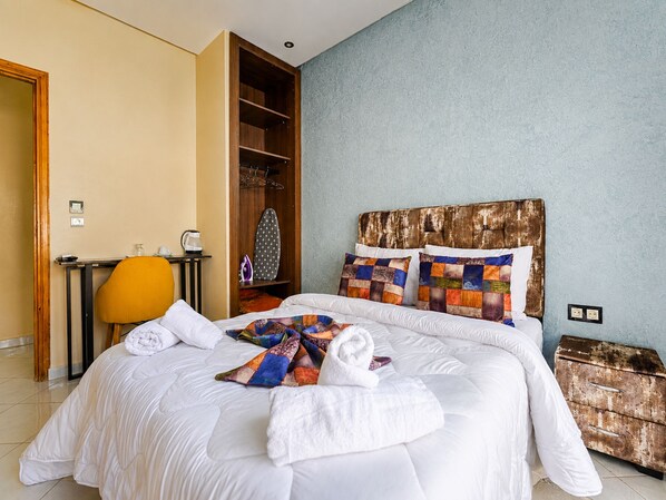 A hotel room with a large bed, a wooden headboard, a small table with a kettle, a chair, and a wardrobe.