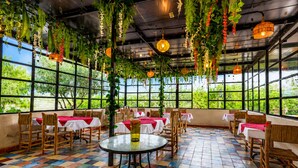 Ein Restaurant mit einem farbenfrohen Fliesenboden, Tischen mit roten Tischdecken, hängenden Pflanzen und großen Fenstern mit Blick auf Grünflächen.