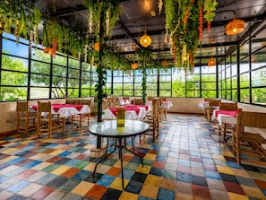 Un restaurante con piso de baldosas coloridas, mesas con manteles rojos, plantas colgantes y ventanas amplias con vista a follaje.