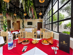 An indoor dining area with tables set for two, featuring a red tablecloth, traditional pottery dishes, and a bottle of water.