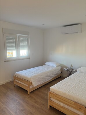 Ein Zimmer mit zwei Holzbett, einem Fenster mit Jalousien, einem kleinen Tisch mit einer Laterne und einer Klimaanlage.