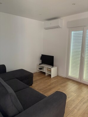 Ein modernes Wohnzimmer mit einem schwarzen Ecksofa, einer weißen Unterhaltungseinheit mit Flachbildfernseher und einem großen Fenster mit Jalousien.