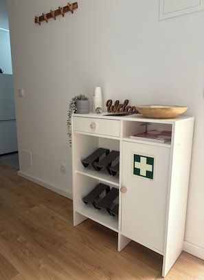 Ein weißer Lagerraum mit einem Holzschild mit der Aufschrift 'Welcome', einem kleinen Topf mit Pflanze und einem Erste-Hilfe-Symbol.