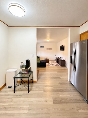 A modern kitchen with a stainless steel refrigerator, a glass-top table with various items, and a wooden floor.