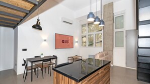 A modern kitchen with a central island, dining area with chairs, pendant lighting, and a large window with blinds. - GuestReady - Apartment with Mezzanine in Benicalap (Valencia)