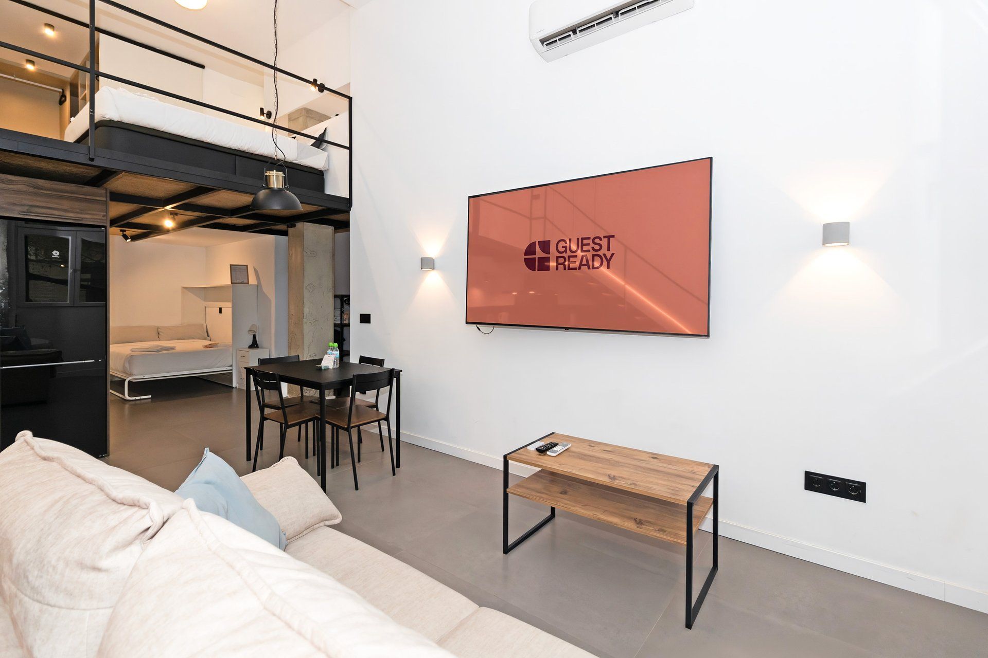 Una habitación de hotel moderna con una cama litera, un escritorio y un sofá. Hay un cartel en la pared que dice 'GUEST READY'.