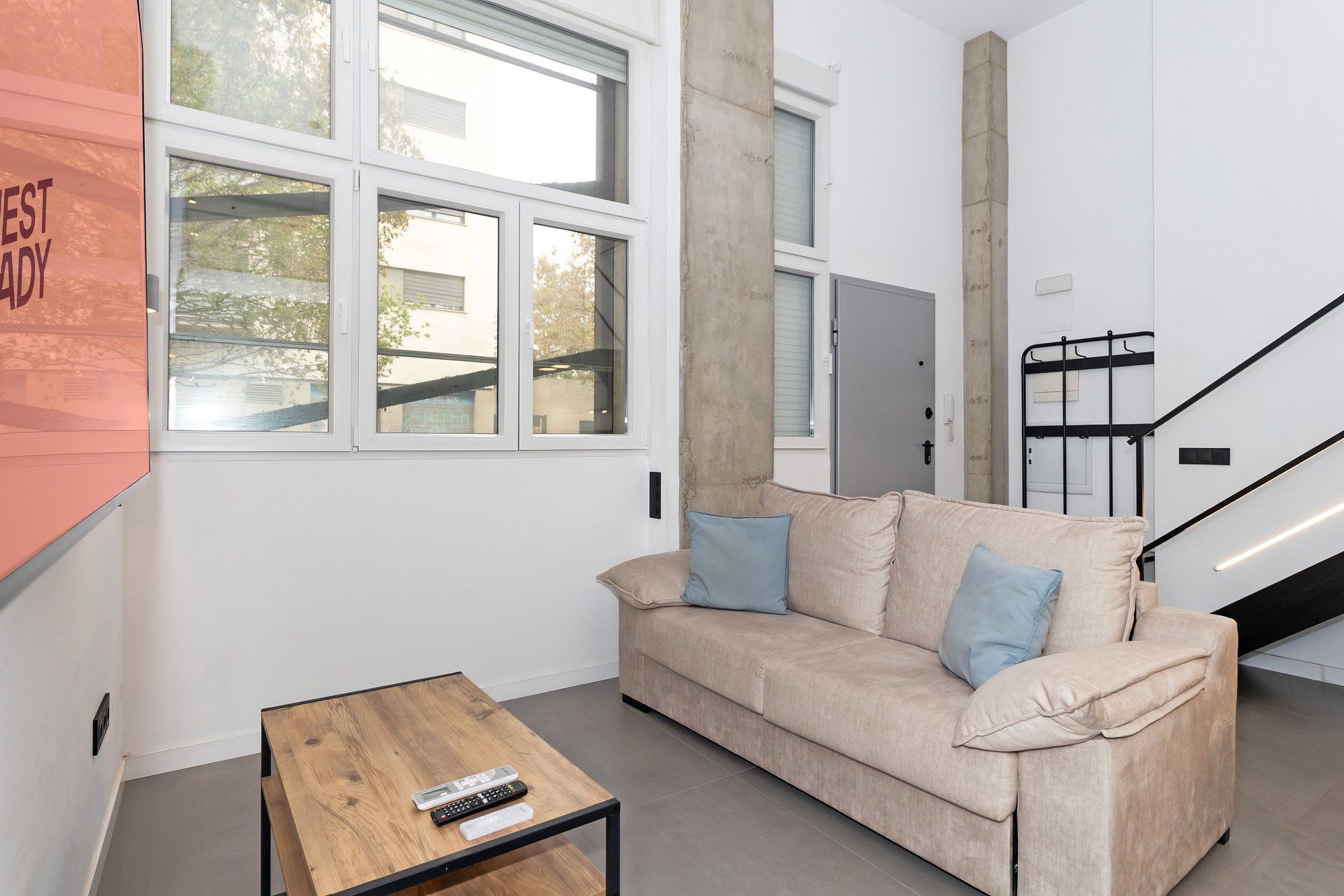 Una sala de estar moderna con un sofá color beige, dos cojines azules, una mesa de centro de madera y una escalera de metal negro.