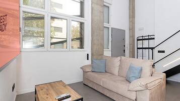 Una sala de estar moderna con un sofá color beige, dos cojines azules, una mesa de centro de madera y una escalera de metal negro.