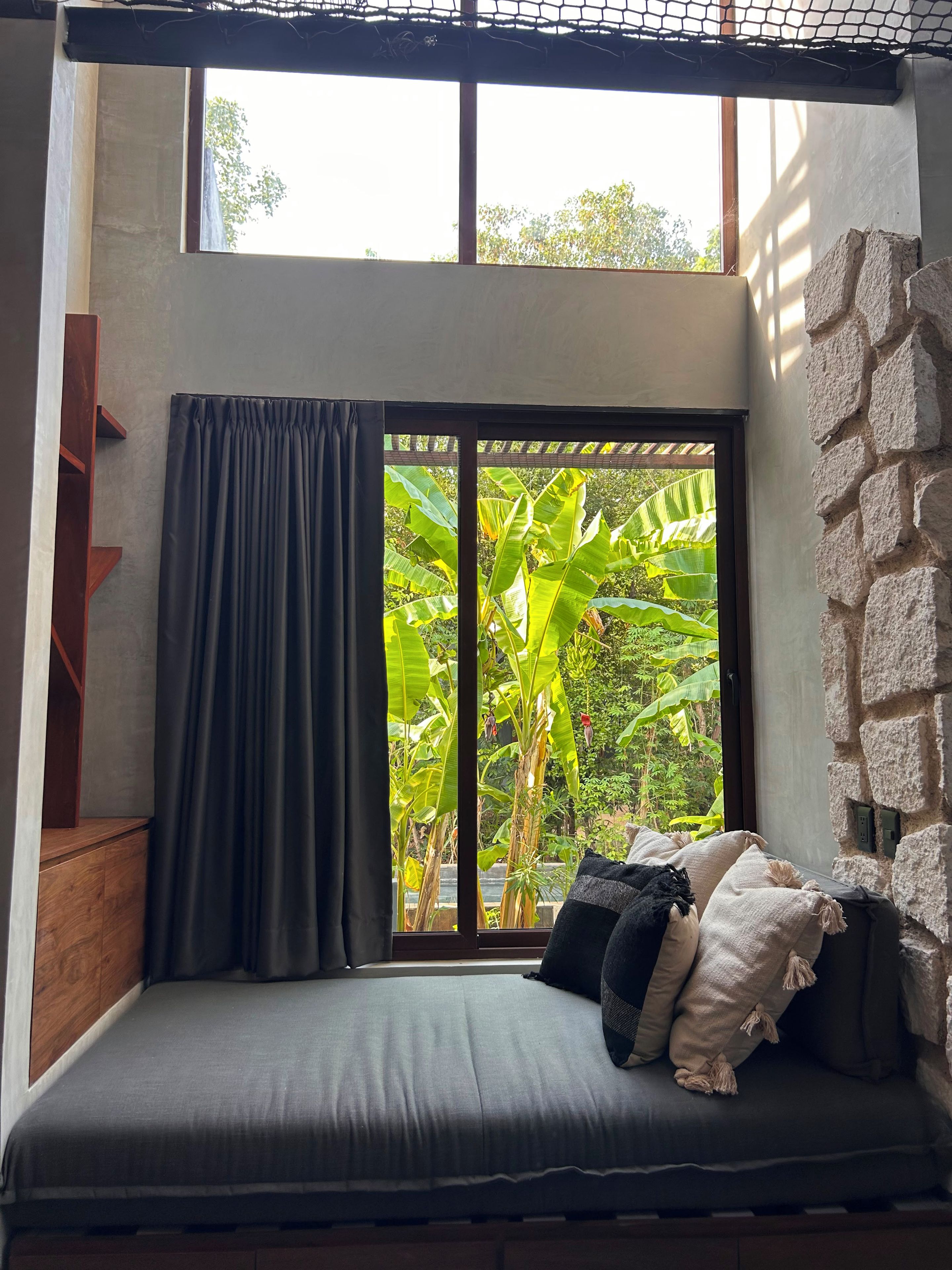 A room with a bed, a window with curtains, a stone wall, and a view of greenery outside.
