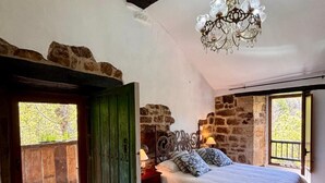 A bedroom with a wooden floor, a bed with a white duvet, a stone wall, a chandelier, and a window with a view of trees. - Lo Macario Pasiego Cabin (Resconorio)