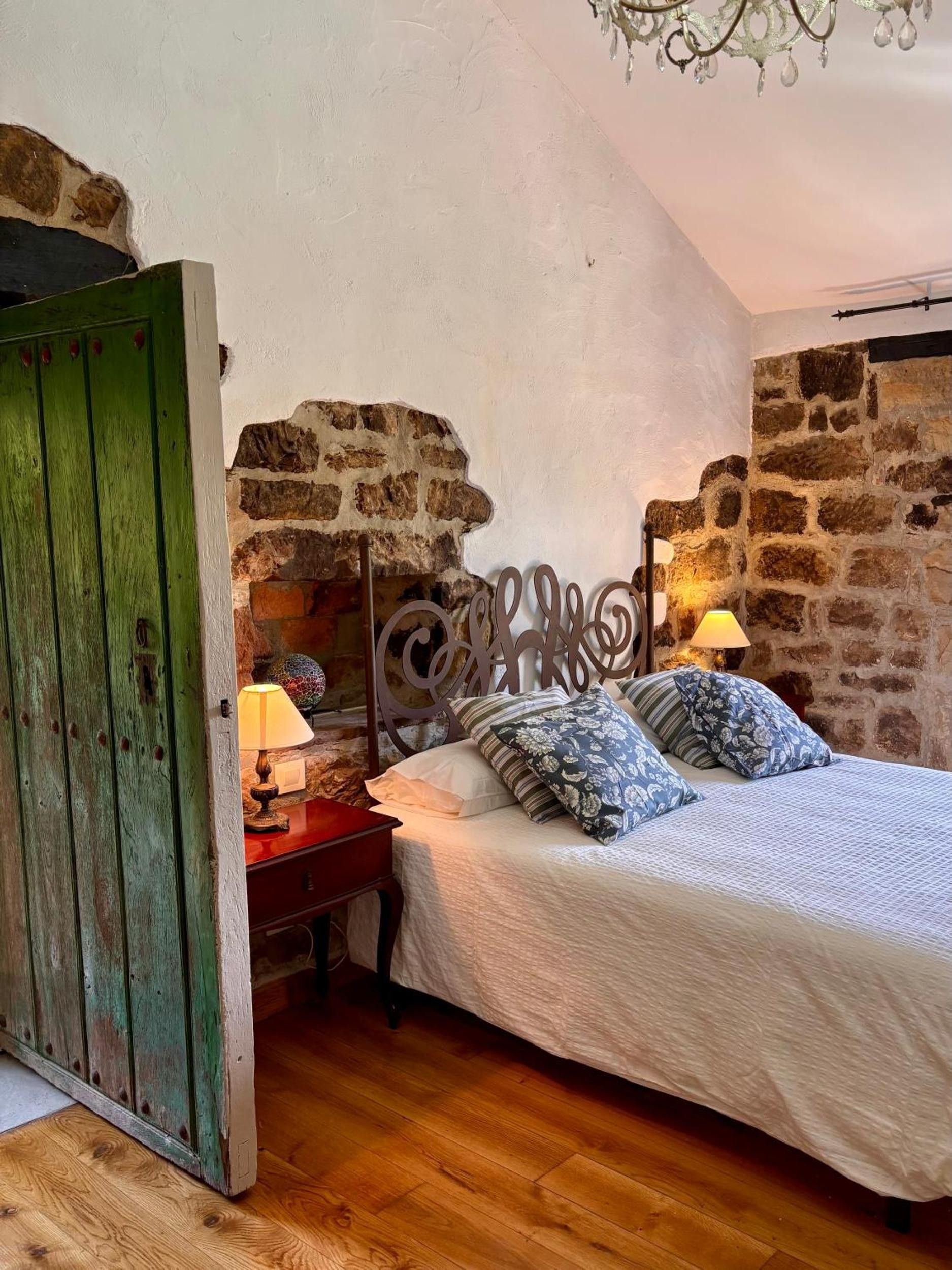 Un dormitorio con pared de piedra, una cama con almohadillas estampadas, una mesita de noche con lámpara y una puerta verde.