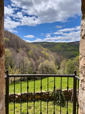 A stone wall with a metal railing, a grassy field, and a hilly landscape with trees under a partly cloudy sky.