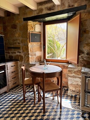 A stone-walled kitchen with a round table, chairs, and a window with a view of trees.
