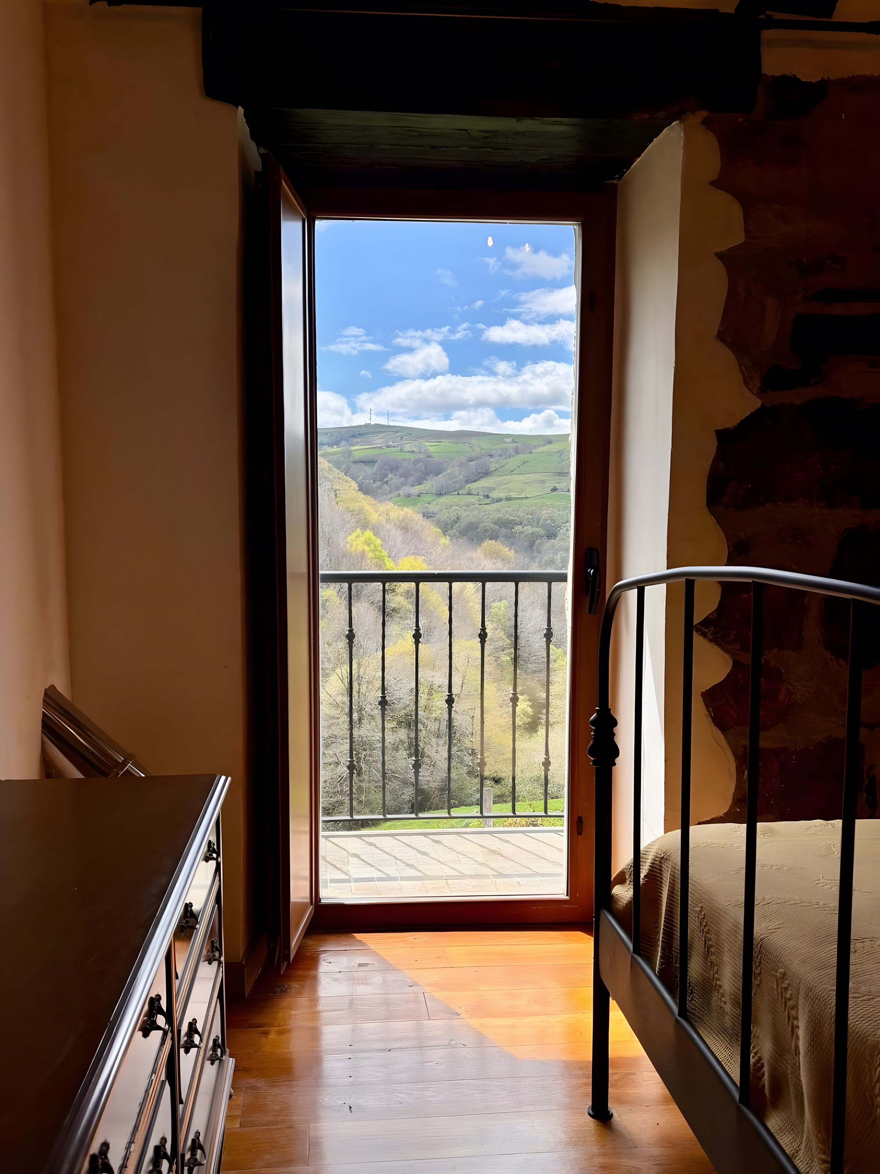 Une pièce avec un plancher en bois, un lit avec un cadre en métal, une commode et une porte donnant sur un balcon avec vue sur un paysage vallonné.