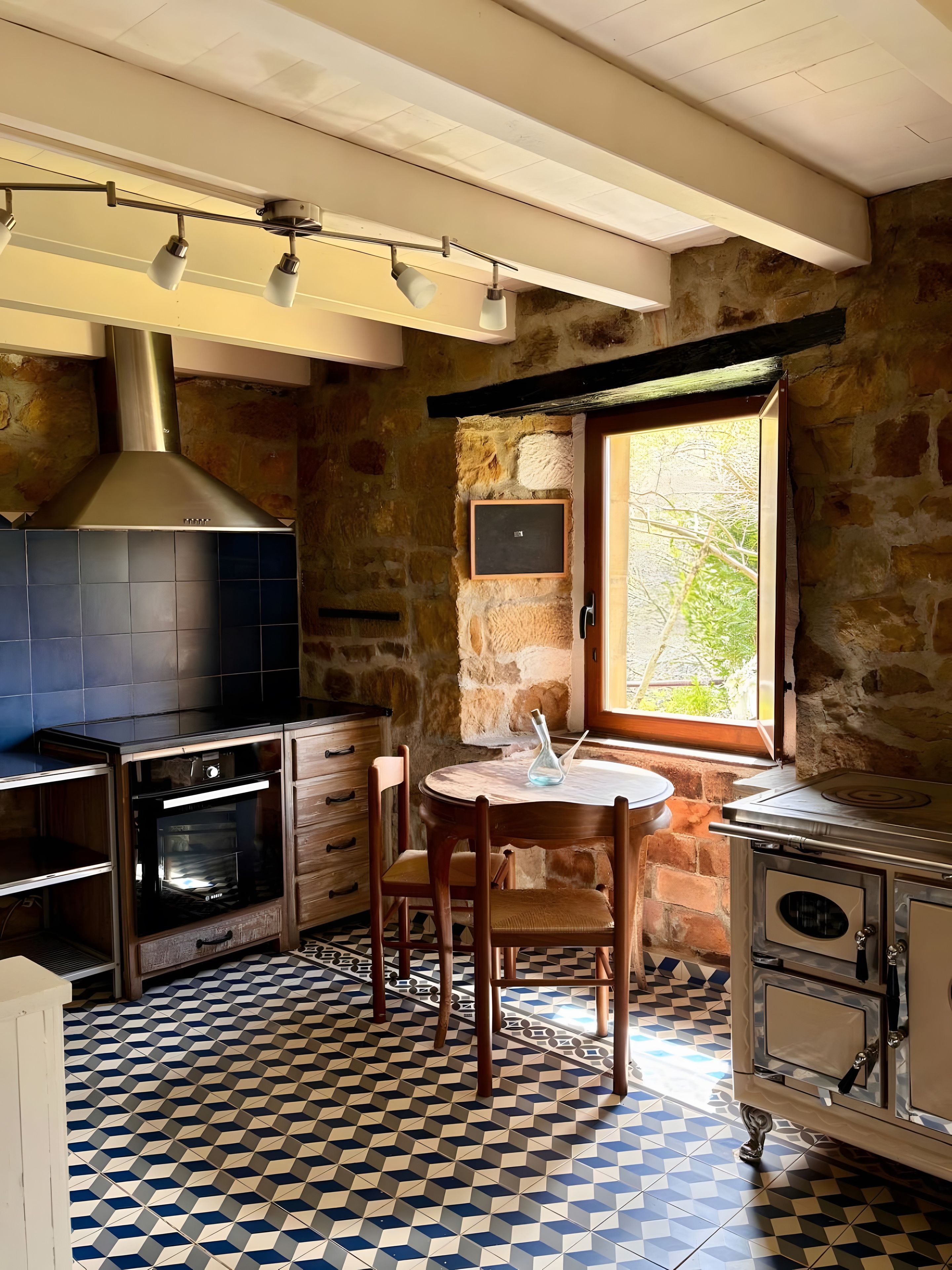 Une cuisine avec un mur en pierre, une fenêtre, une table ronde en bois et un four encastré.