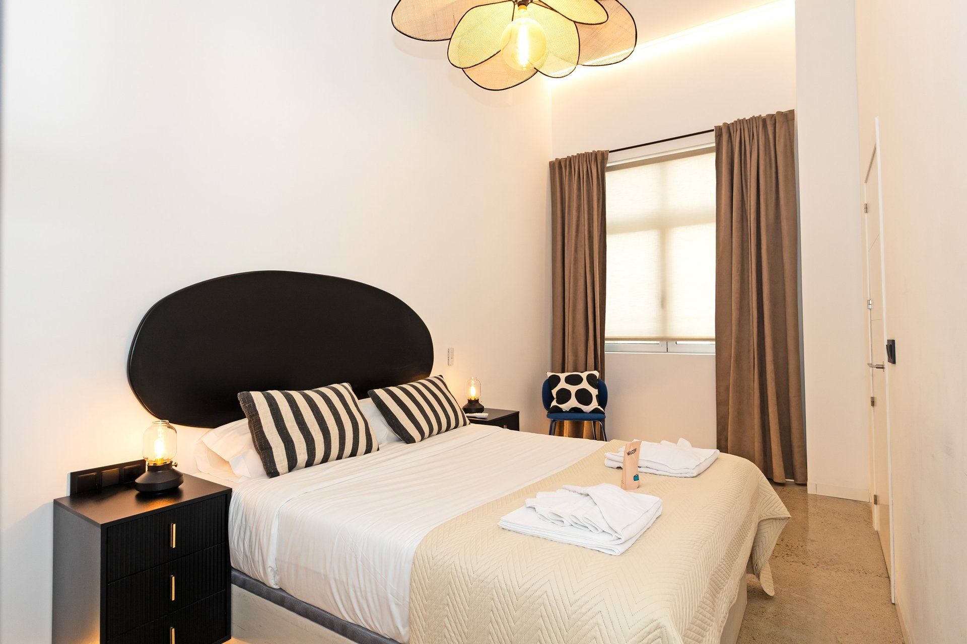 Una habitación de hotel con cama, mesita de noche, lámpara, sillón y ventana con cortinas.