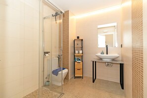 Un baño con ducha envuelta en vidrio, inodoro, lavamanos y estante con artículos de aseo.