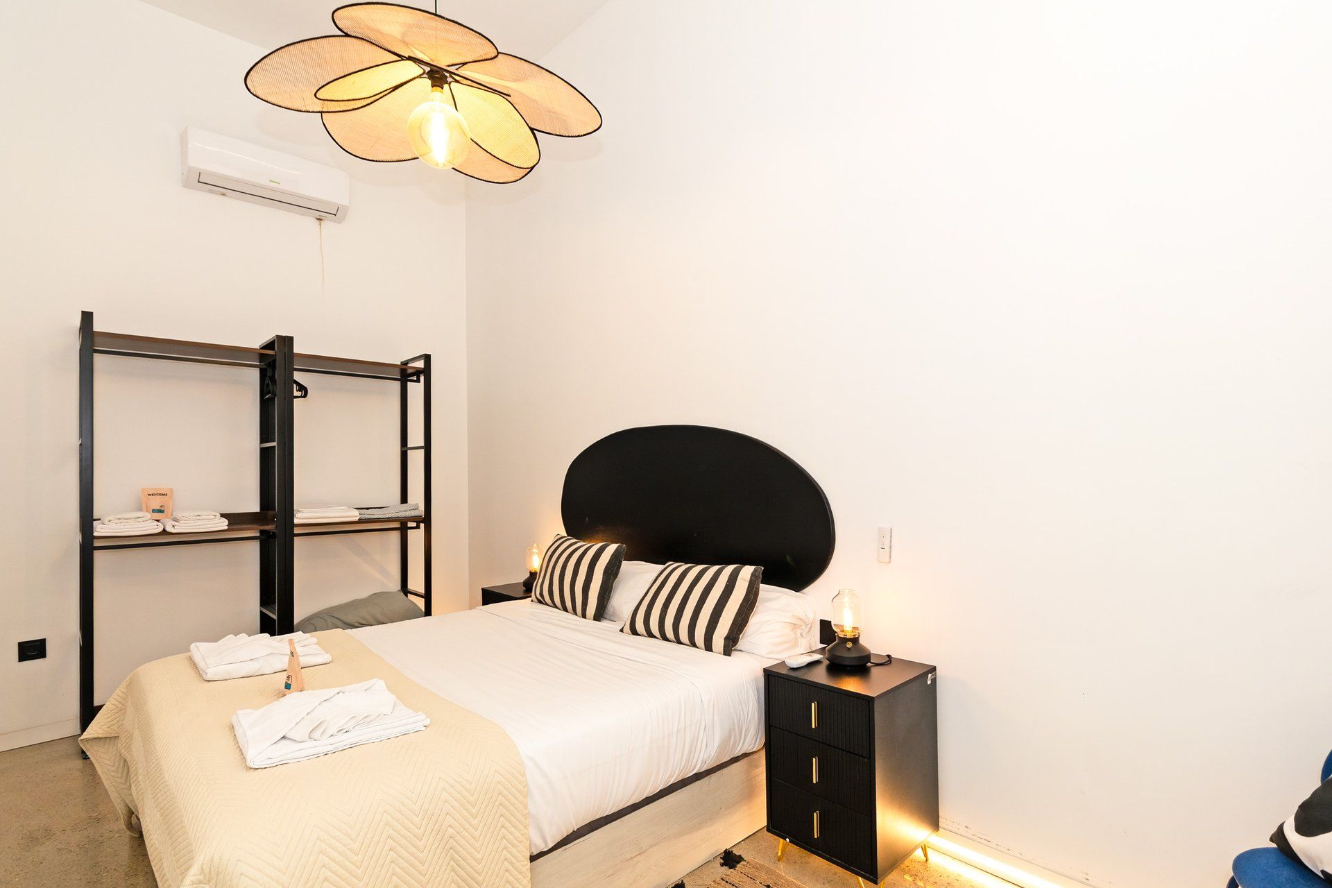 Un dormitorio con cama, mesita de noche con lámpara, aire acondicionado de pared y ventilador de techo.