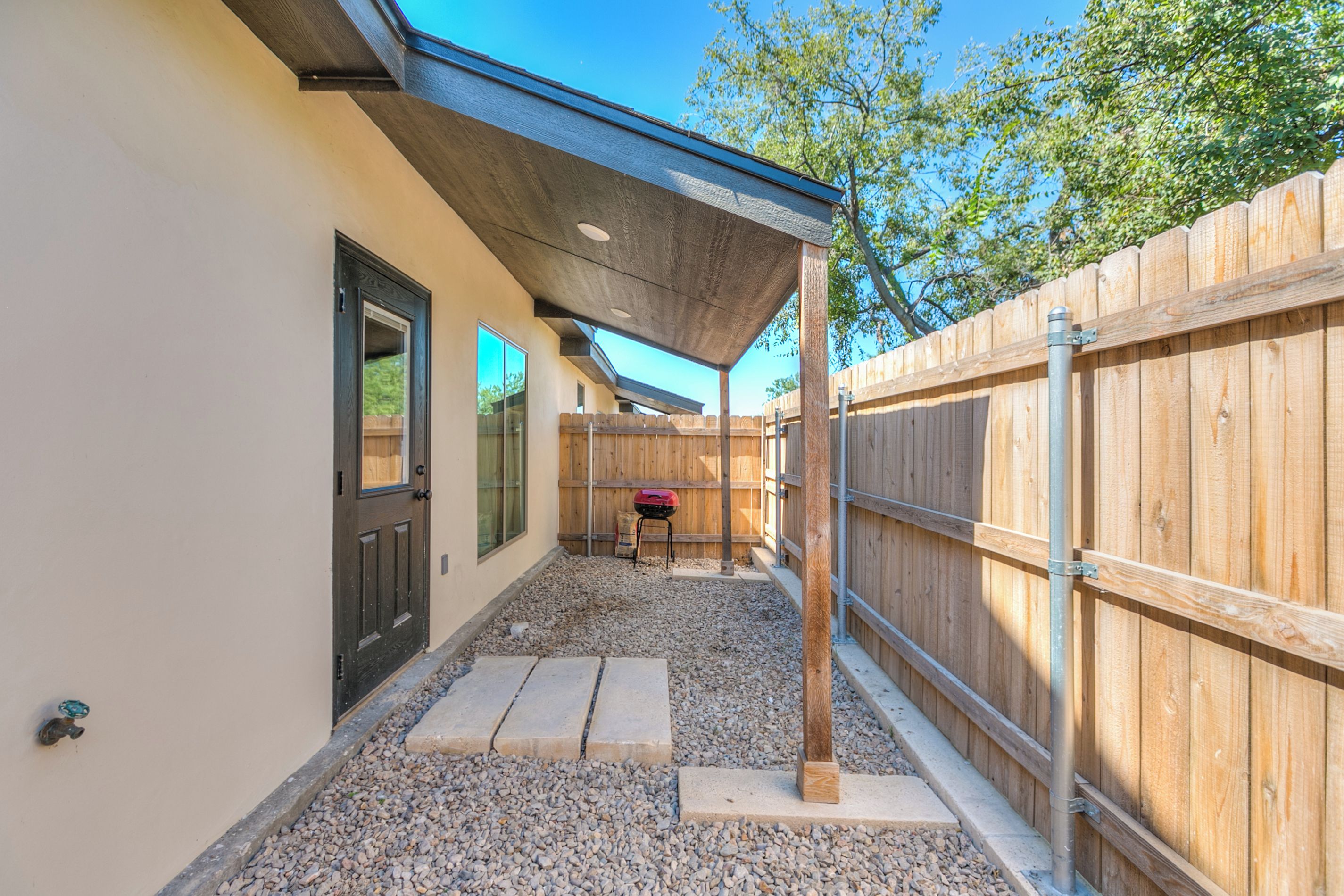 A covered outdoor patio area with a wooden fence, a barbecue grill, and a gravel pathway.
