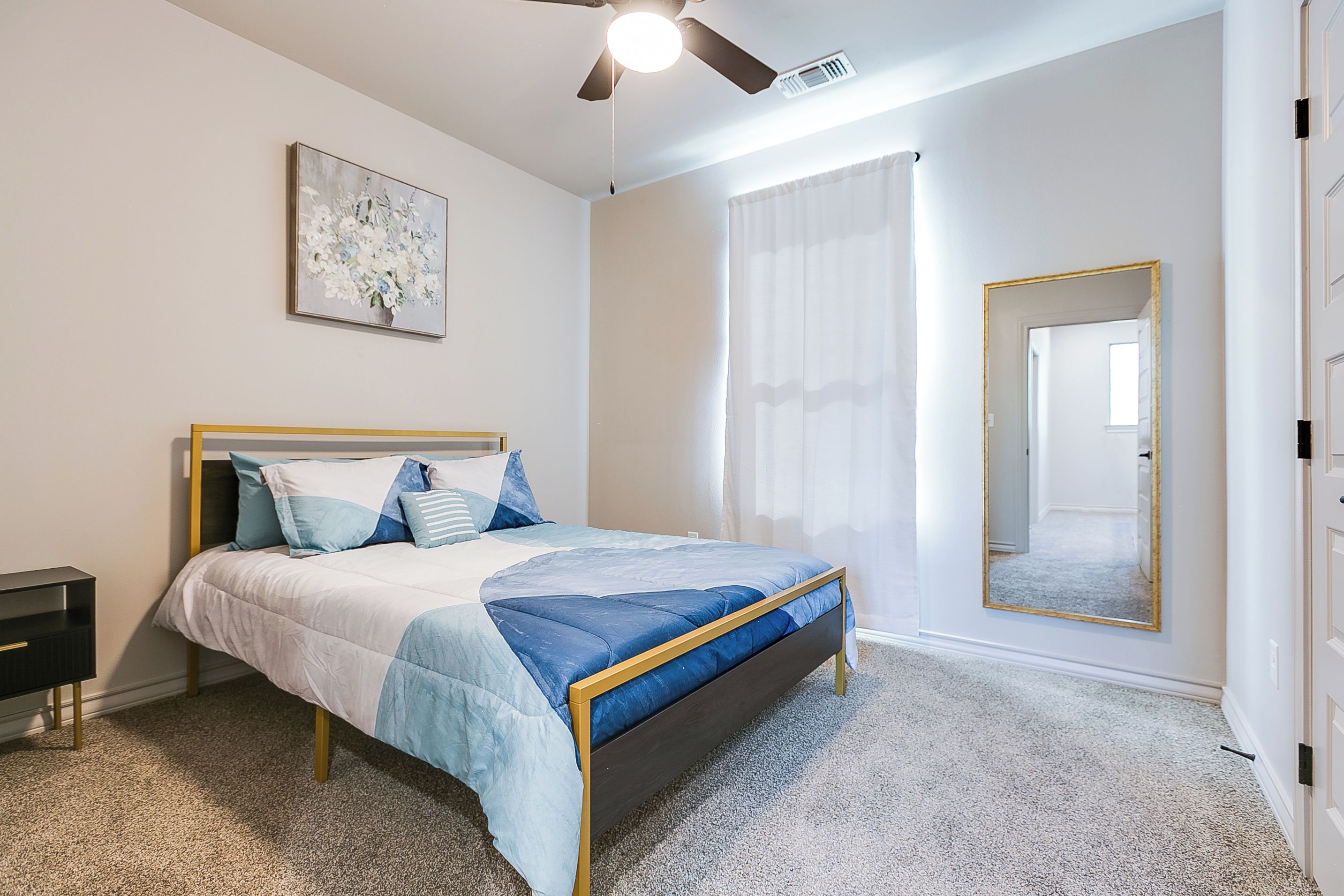 A bedroom with a bed, a nightstand, a ceiling fan, a window with curtains, and a mirror.