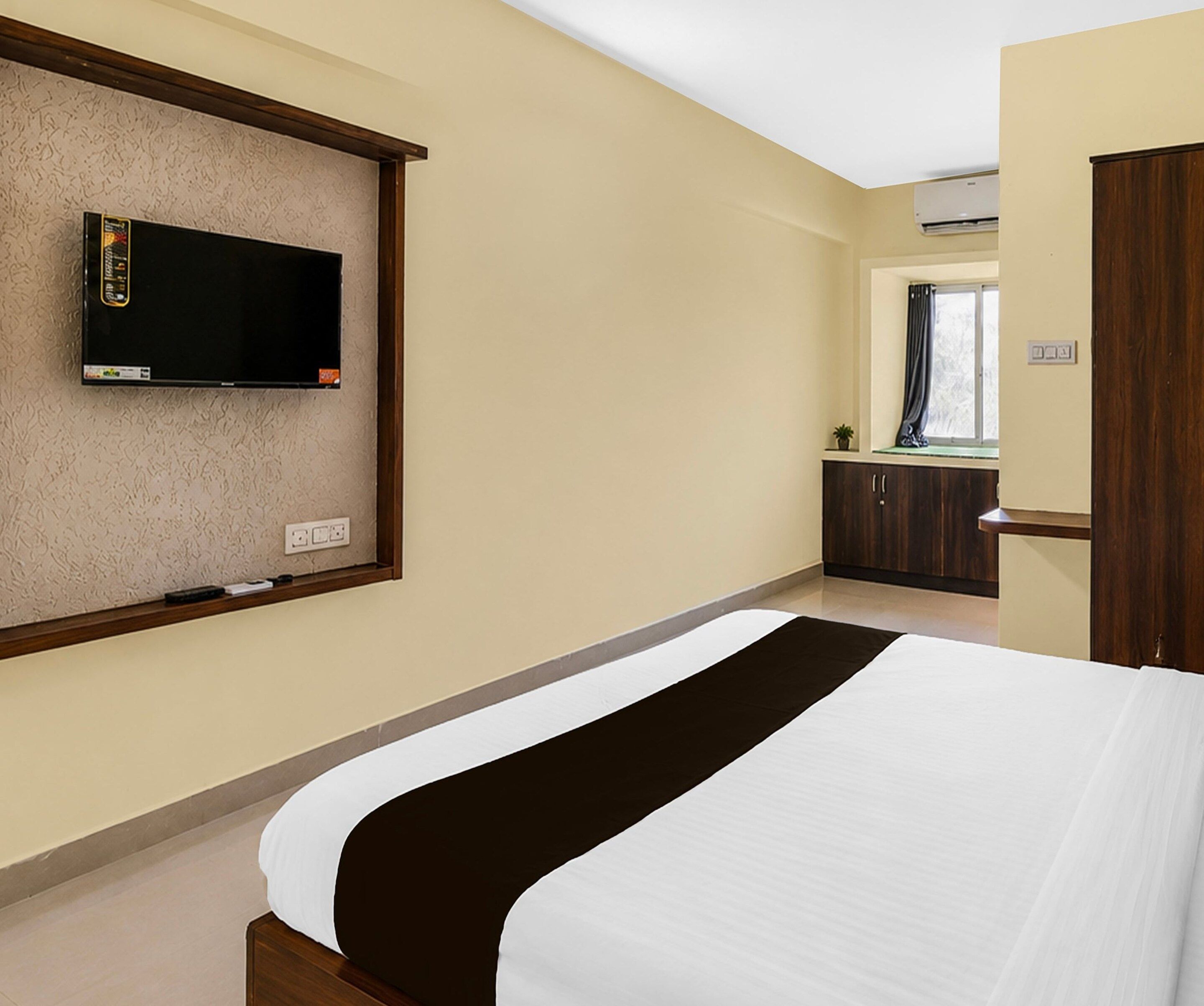 A hotel room with a bed, a television mounted on the wall, a window with curtains, and a wardrobe.