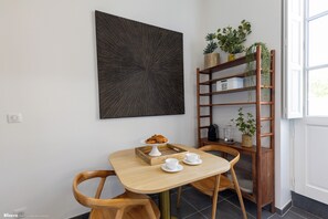 Zona pranzo con tavolo in legno, due sedie, un elemento decorativo a parete, una libreria con piante e una finestra che lascia entrare luce naturale.