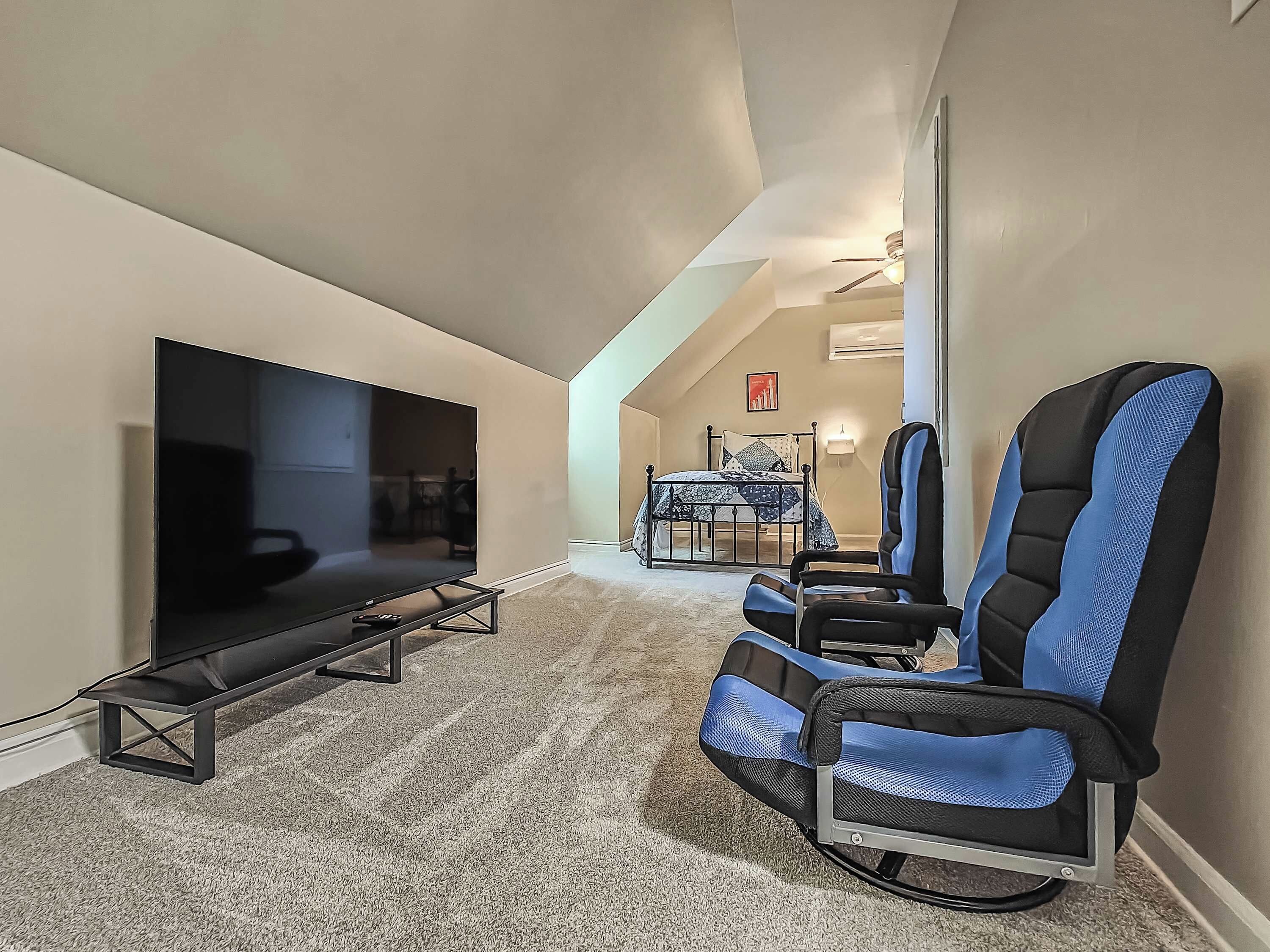 A modern living room with a flat-screen TV on a stand, a ceiling fan, a bed with a patterned bedspread, and two blue and black office chairs.