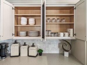 Una cocina con alacenas abiertas que contienen platos, tazones y cristalería. Hay tres recipientes etiquetados como café, leche y azúcar. Una pequeña planta en maceta está sobre la encimera.