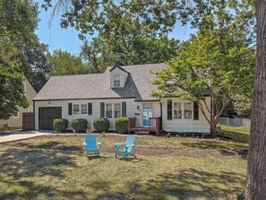 A two-story house with a garage, a covered porch, and two blue Adirondack chairs in the front yard. - Charming 4 BDRM New Listing in OP! (Overland Park)