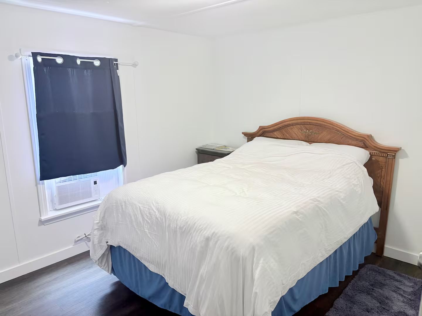 A bedroom with a bed, a window with a curtain, a nightstand, and a wooden headboard.