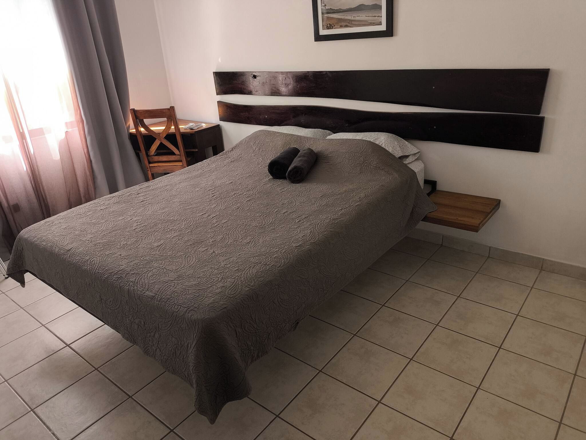 Ein Hotelzimmer mit einem Bett, einem Nachttisch aus Holz, einem Sessel, einem kleinen Tisch und einem gerahmten Bild an der Wand.