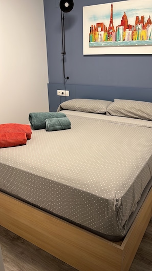 A bed with a polka dot bedspread, two rolled towels, a lamp, and a painting of a cityscape on the wall. - Arenal Beach, Bol, Calpe Lookout (Calpe)