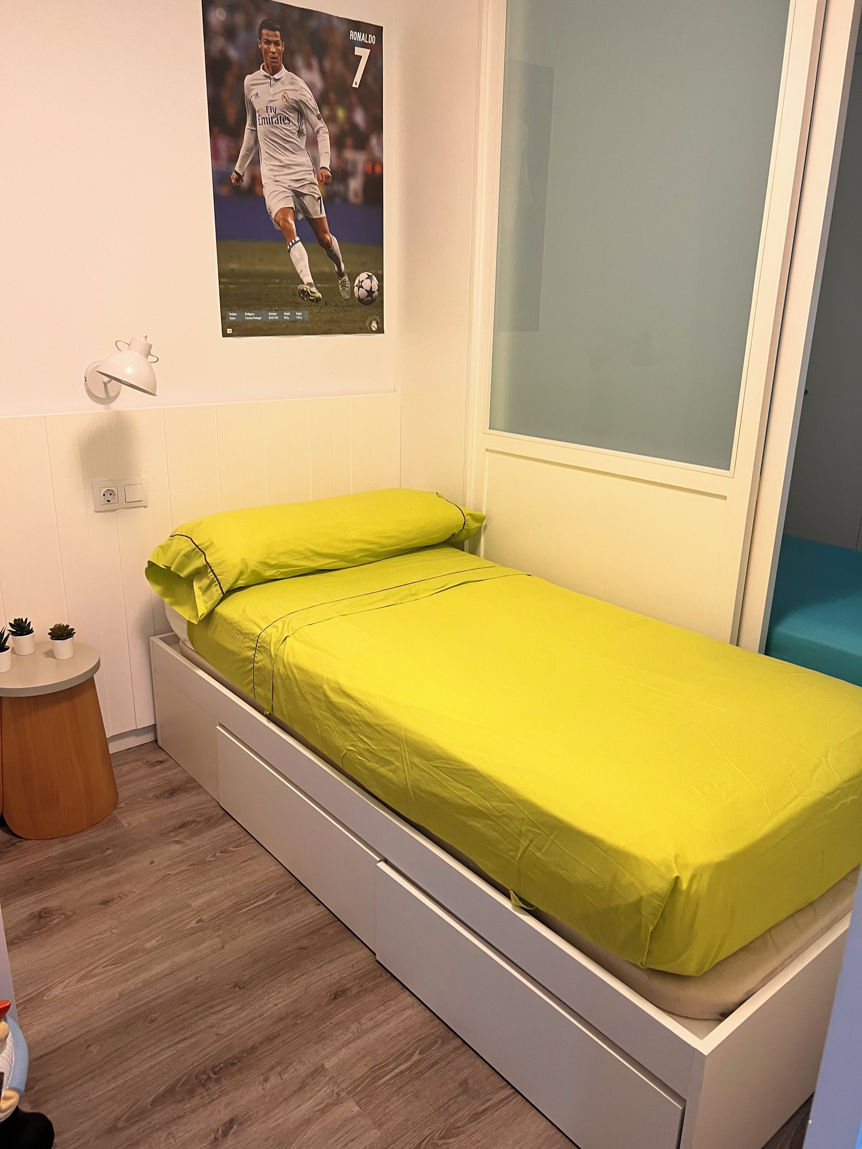 A single-bed room with a green mattress, a poster of a soccer player, a bedside table with a lamp, and a window with a view.