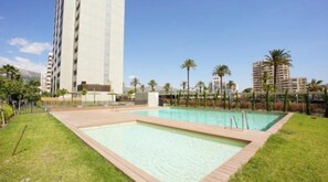 A swimming pool with a wooden deck, surrounded by a fence and palm trees, with high-rise buildings in the background. - Arenal Beach, Bol, Calpe Lookout (Calpe)