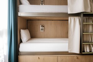 Ein Etagenbett mit einem Bett darunter, eine kleine Ablage mit Steckdose und ein Vorhang auf der rechten Seite.