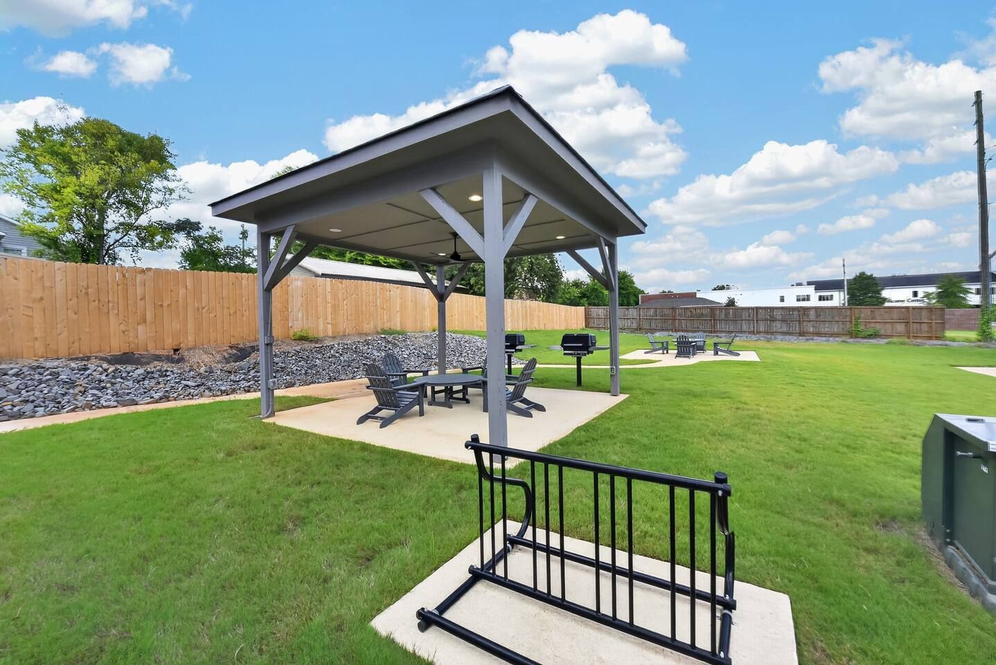 A covered outdoor seating area with a grill and chairs, surrounded by a wooden fence and a grassy lawn.