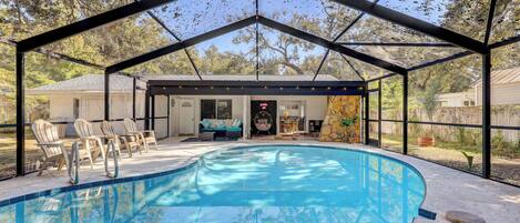 A covered swimming pool with a glass ceiling, surrounded by outdoor furniture and a house in the background.